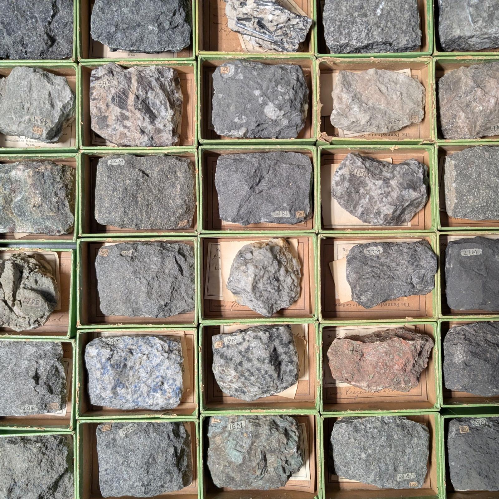 A gridded assortment of grey, pink, and brown rock specimens in a green metal drawer